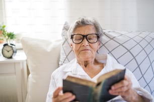 안경을 쓴 아픈 노파가 집이나 병원의 침대에 누워 성경을 읽고 있다.