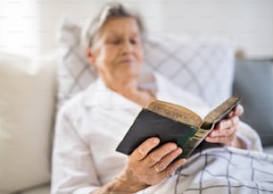 A sick senior woman lying in bed at home or in hospital, reading bible book.