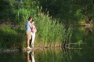 Happy young pregnant couple hugging in nature, reflecting in the lake