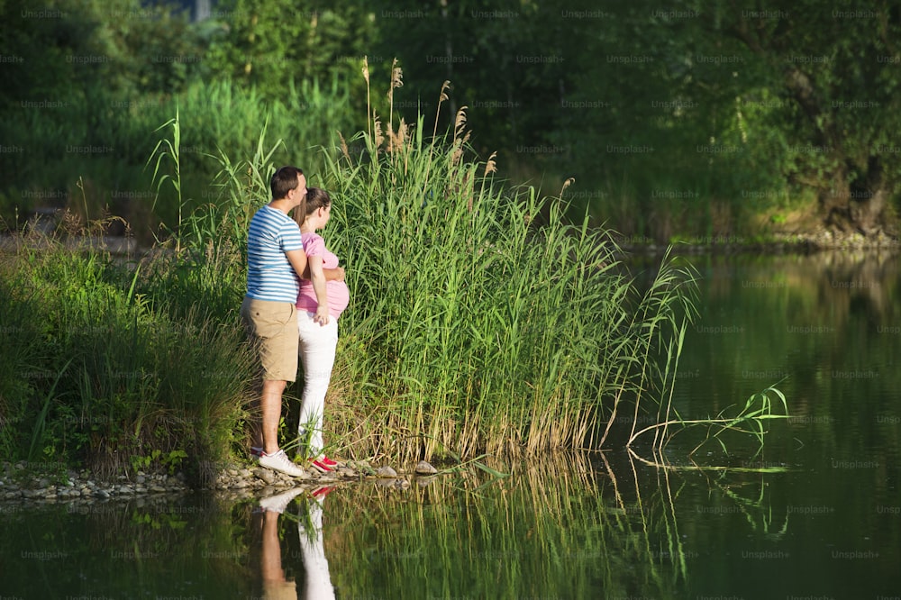 Feliz joven pareja embarazada abrazándose en la naturaleza, reflexionando en el lago