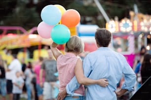 Senior couple having a good time at the fun fair. Woman holding colorful balloons. Sunny summer day. Rear view.