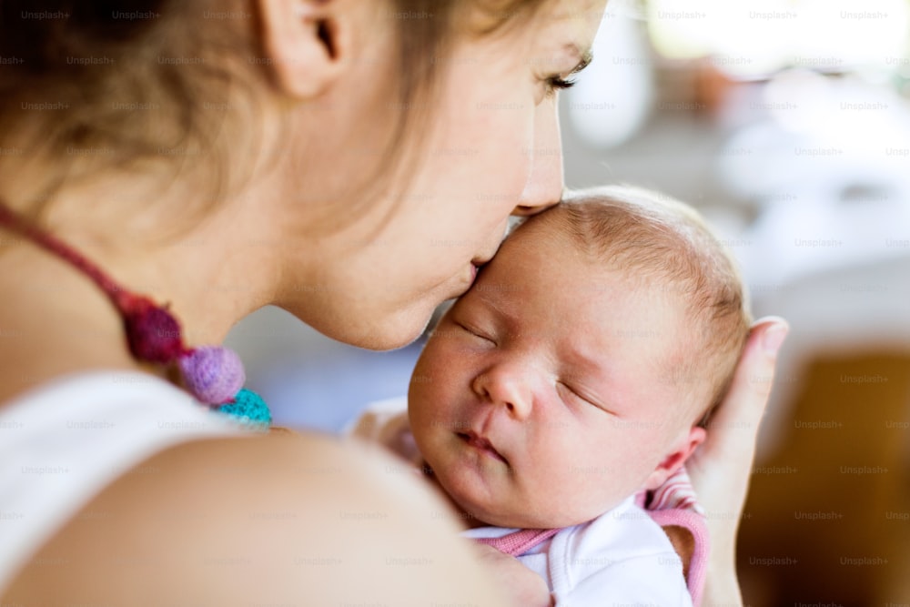 家でかわいい生まれたばかりの女の赤ちゃんを抱きしめ、額にキスをする美しい若い母親。