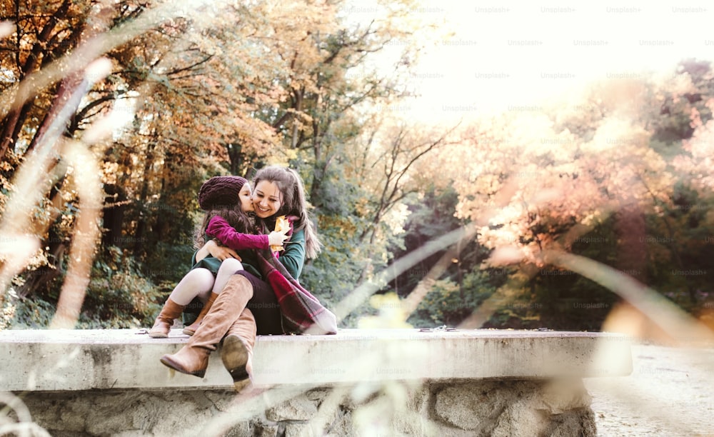 A portrait of young mother with a toddler daughter hugging and kissing in forest in autumn nature. Copy space.
