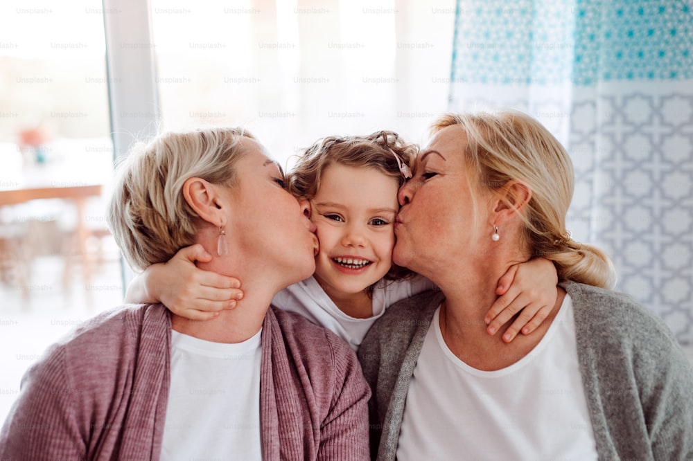 A portrait of happy small girl with mother and grandmother at home, kissing.