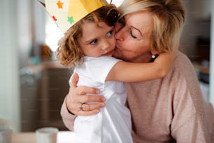 A portrait of small girl and grandmother with paper crown hugging and kissing at home.