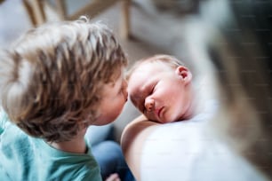 Um menino pequeno beijando um irmãozinho recém-nascido dormindo em casa, uma mãe segurando-o.