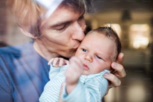 A young father holding a newborn baby at home, kissing. Shot through glass.