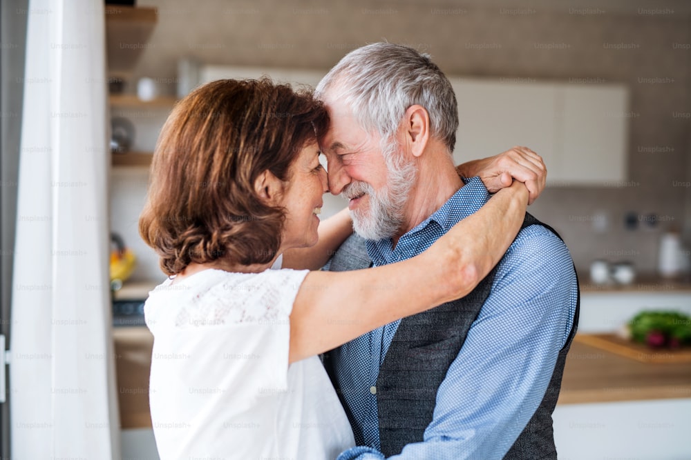 An affectionate senior couple in love standing indoors at home, hugging.