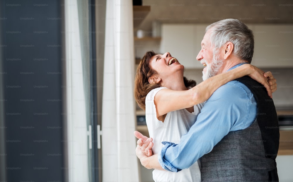 A senior couple standing indoors at home, laughing and hugging.