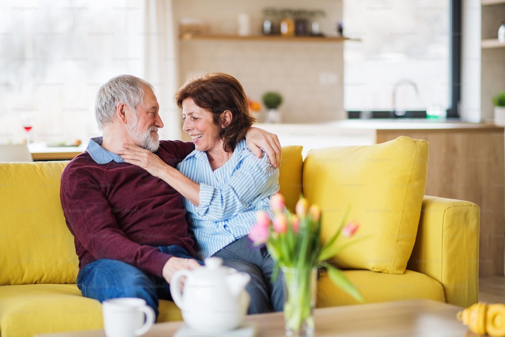 Un couple de personnes âgées amoureuses affectueuses assis sur un canapé à l’intérieur de la maison, en train de parler.