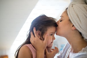 Portrait of mother kissing small daughter indoors in bathroom at home.