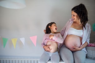 Portrait of happy pregnant woman with small daughter indoors at home, showing belly.