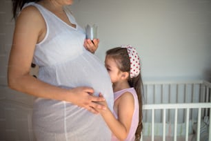 Side view of small girl kissing belly of unrecognizable pregnant mother indoors at home.