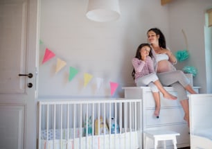 Porträt einer glücklichen schwangeren Frau mit kleiner Tochter drinnen zu Hause, auf Kommode sitzend.