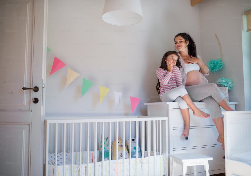 Porträt einer glücklichen schwangeren Frau mit kleiner Tochter drinnen zu Hause, auf Kommode sitzend.