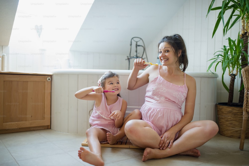Retrato da mulher grávida feliz com a filha pequena dentro de casa no banheiro em casa, escovando os dentes.