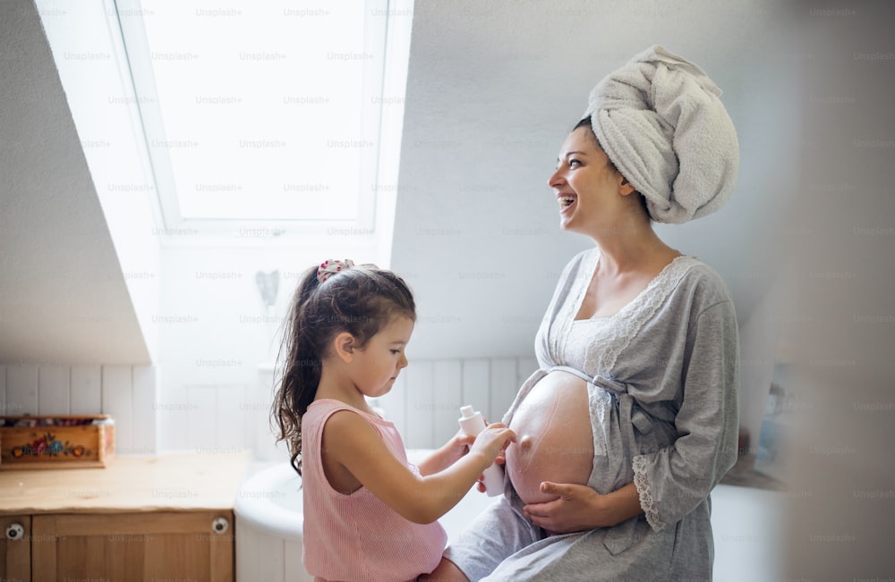 Happy petite fille crémant le ventre de la mère enceinte à l’intérieur dans la salle de bain à la maison.
