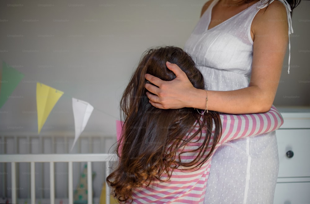 Porträt einer nicht wiederzuerkennenden schwangeren Frau mit kleiner Tochter drinnen zu Hause, umarmend.