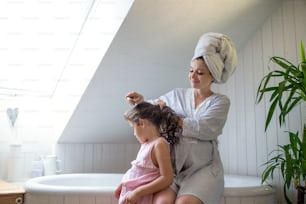 Side view portrait of pregnant woman with small daughter indoors in bathroom at home, brushing hair.