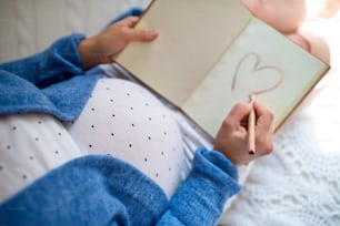 Draufsicht auf eine nicht wiederzuerkennende schwangere Frau in Innenräumen zu Hause, die Herzform in Notizbuch zeichnet.