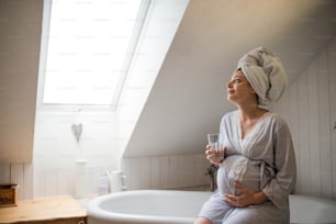 Portrait of happy pregnant woman indoors in bathroom at home, holding glass of water. Copy space.