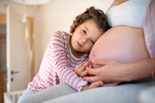 Portrait of small girl hugging belly of unrecognizable pregnant mother indoors at home.
