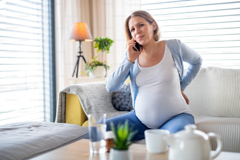 A portrait of pregnant woman in pain indoors at home, making phone call.
