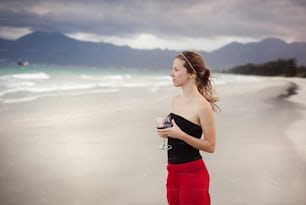 赤ワインのグラスを持つ美しい女性がリラックスして砂浜で海の景色を楽しんでいる