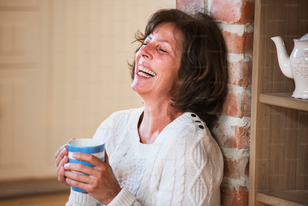 Donna anziana a casa che si gode la bevanda calda, ridendo. Primo piano.