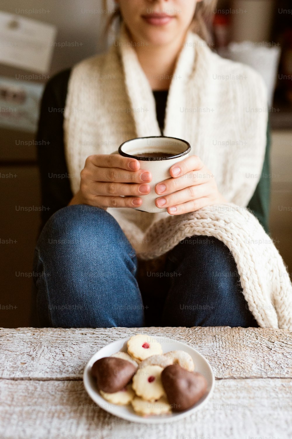 Unkenntliche Frau mit weißem Strickschal, die eine Tasse mit Kaffee hält. Marmeladenplätzchen und Lebkuchen auf einem alten Holztisch.