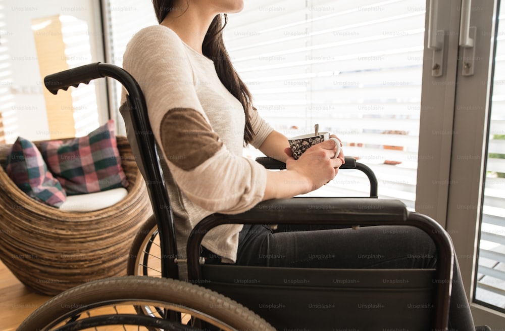 Giovane donna disabile irriconoscibile in sedia a rotelle a casa nel suo salotto, guardando fuori dalla finestra, con in mano una tazza di tè o caffè