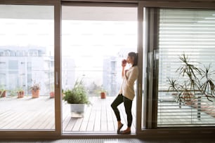 Beautiful young woman relaxing on balcony with city view holding cup of coffee or tea, drinking from it