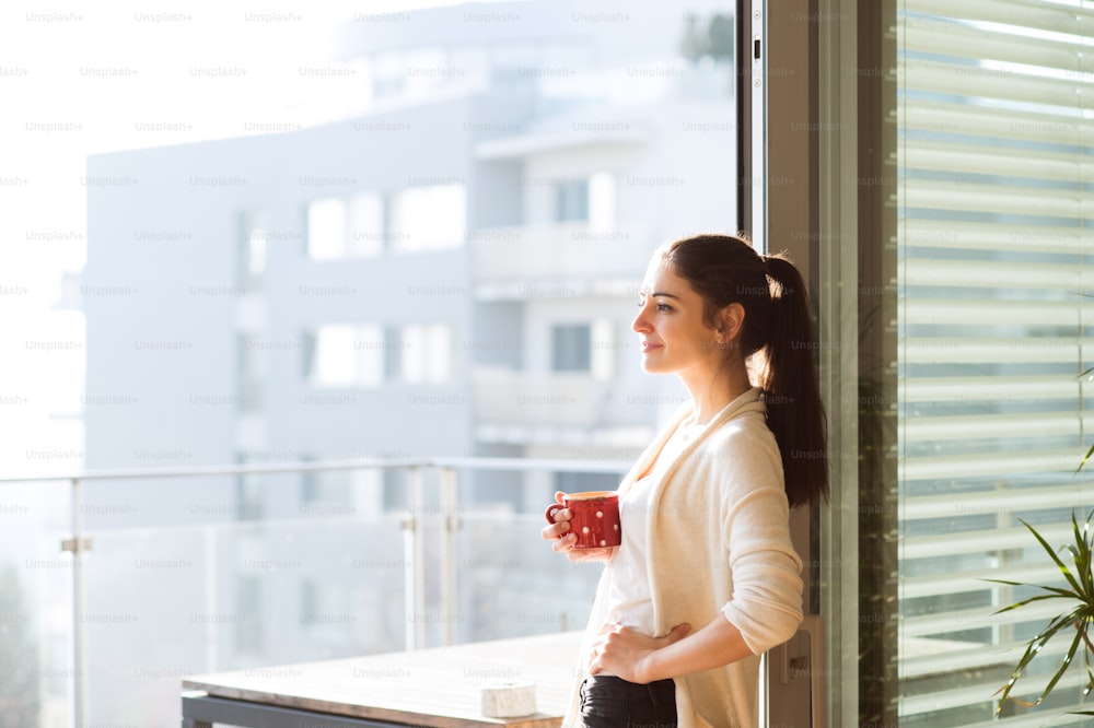 Hermosa joven relajándose en el balcón con vista a la ciudad sosteniendo una taza de café o té