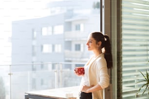 Mulher jovem bonita que relaxa na varanda com vista da cidade segurando a xícara de café ou chá
