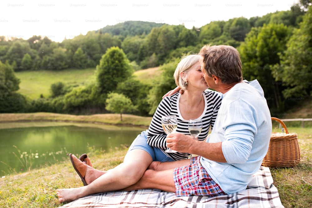 Beau couple de personnes âgées au lac en train de pique-, assis sur une couverture, buvant du vin et s’embrassant.