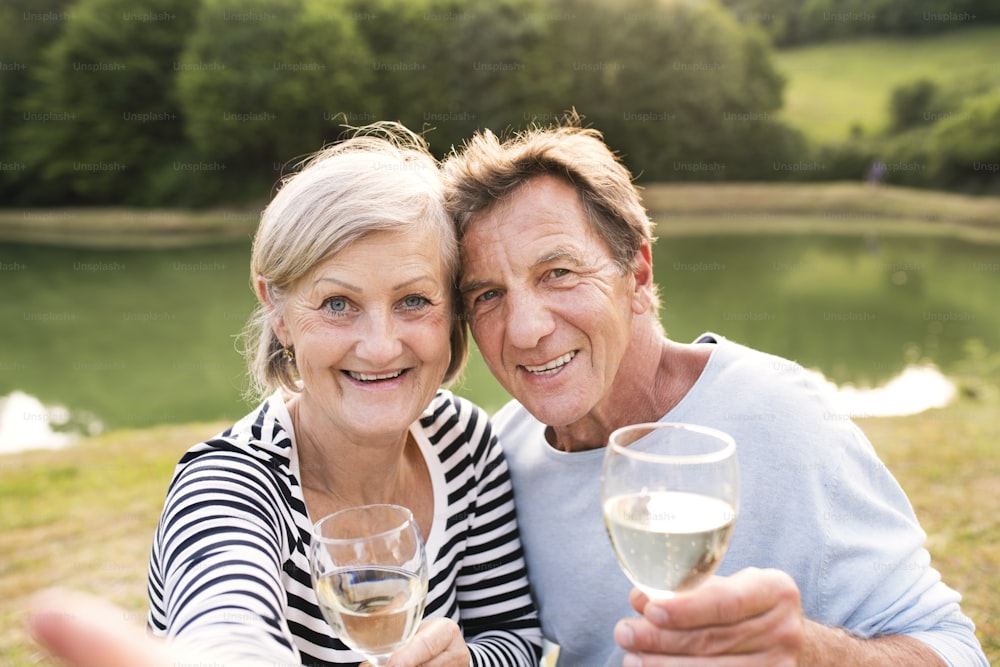 Bella coppia anziana al lago che fa un picnic, seduta sulla coperta, bevendo vino.