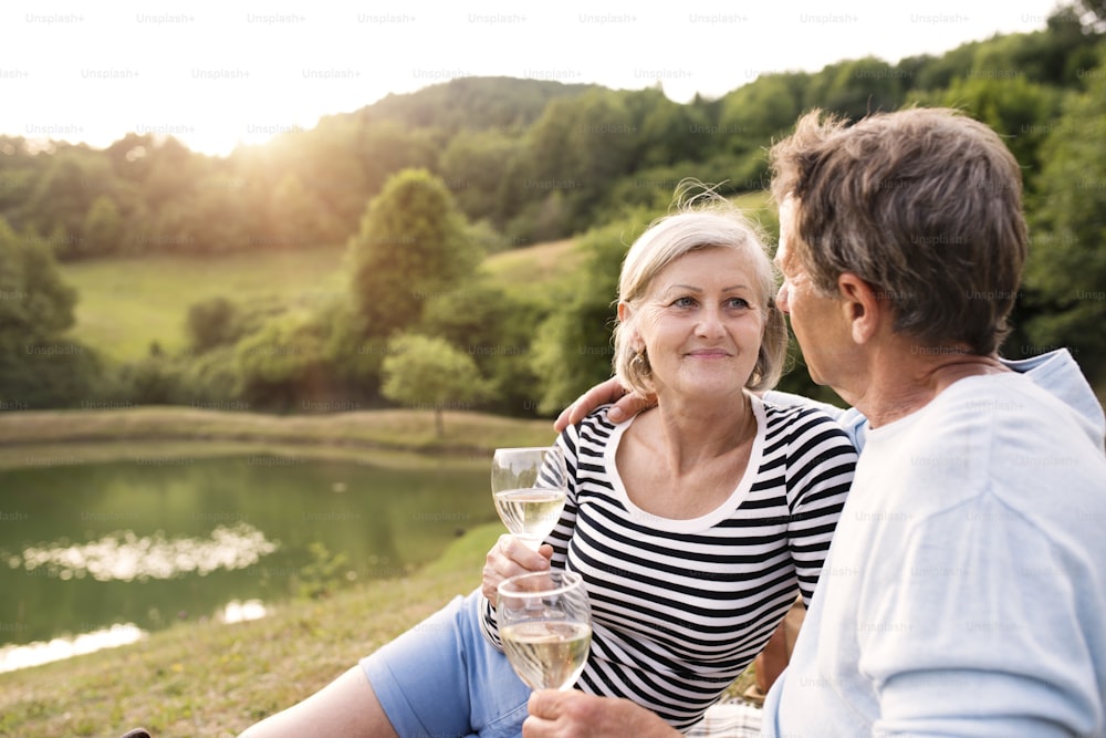 Bella coppia anziana al lago che fa un picnic, seduta sulla coperta, bevendo vino.