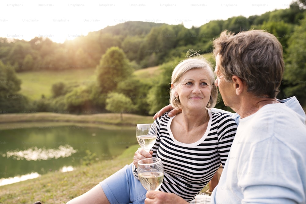 Beautiful senior couple at the lake having a picnic, sitting on blanket, drinking wine.