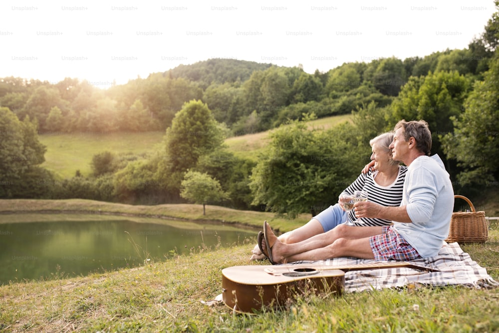 Bella coppia anziana al lago che fa un picnic, seduta sulla coperta, bevendo vino.