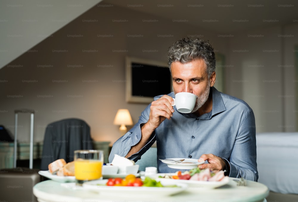 Mûr, bel homme d’affaires prenant le petit déjeuner dans une chambre d’hôtel, buvant du café.