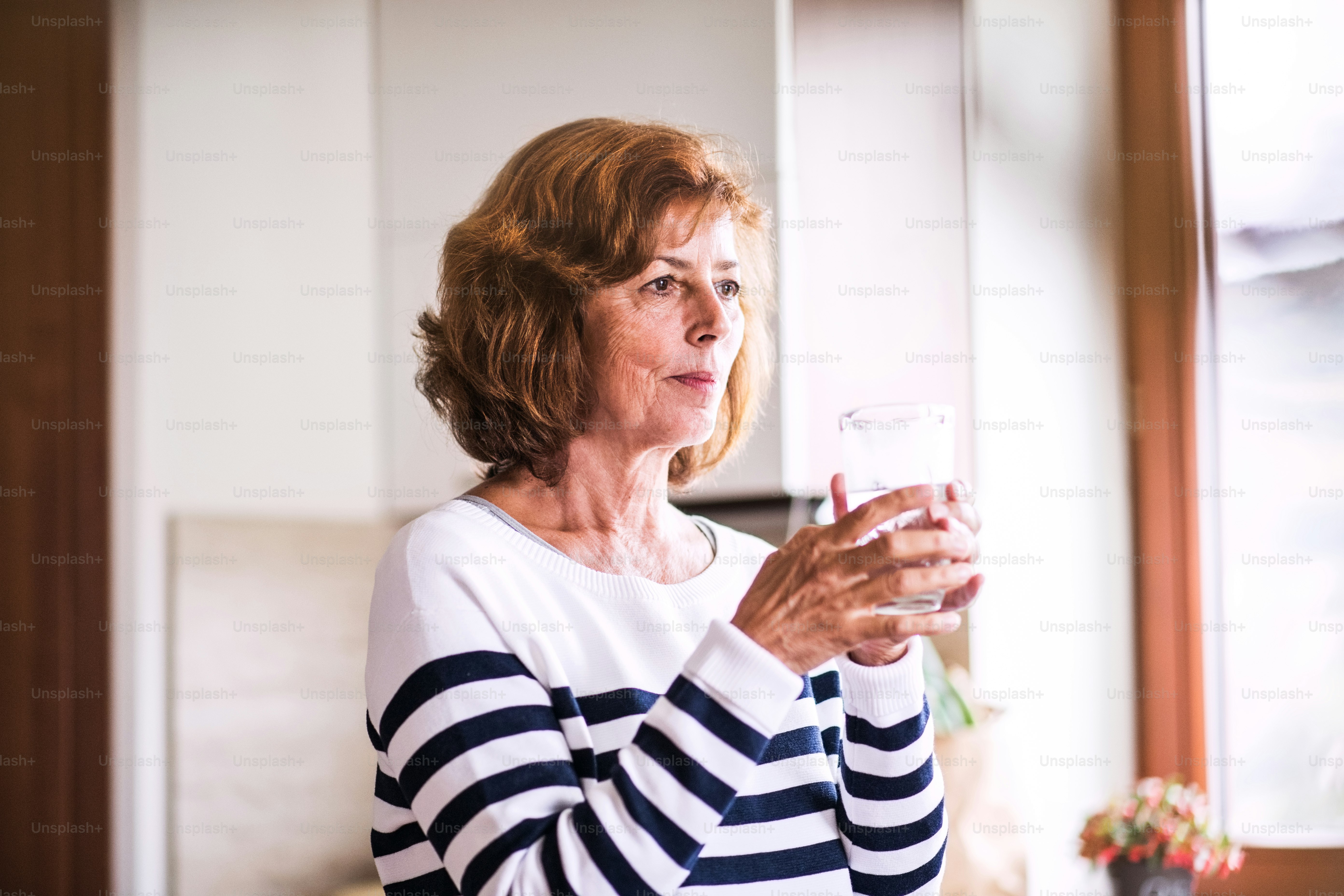 Femme âgée dans la cuisine. Une vieille femme à l’intérieur de la maison, tenant un verre d’eau.