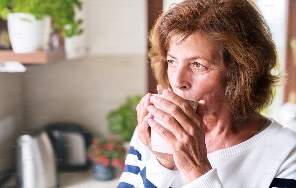 Mulher sênior na cozinha. Uma mulher idosa dentro da casa, segurando e bebendo uma xícara de café.