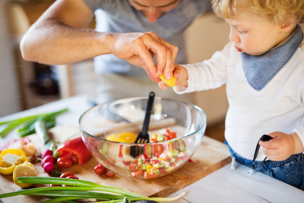 Pai jovem irreconhecível com um menino cozinhando. Um homem com seu filho fazendo salada de legumes.