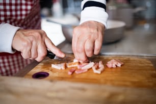 Mujer mayor irreconocible cocinando en casa. Primer plano de las manos cortando tocino.