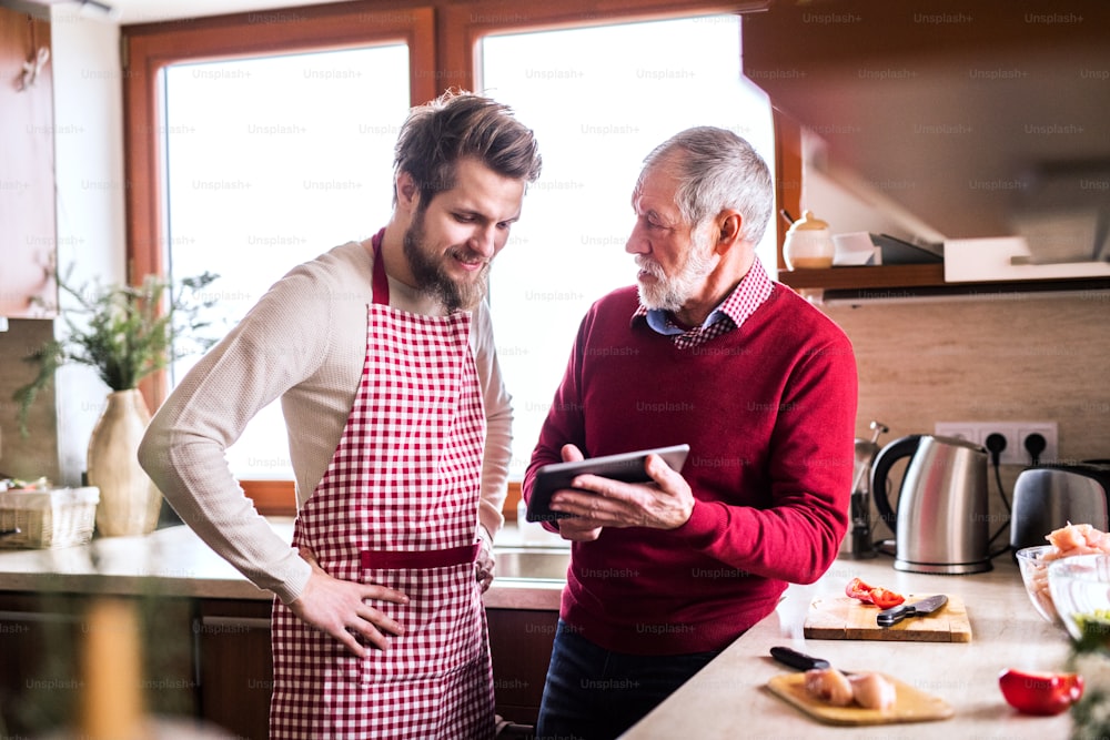 Hipster son with his senior father cooking in the kitchen. Two men with a tablet.