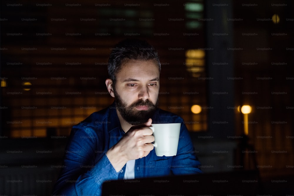 Ein gutaussehender Mann, der nachts zu Hause an einem Laptop arbeitet und eine Tasse Kaffee in der Hand hält.