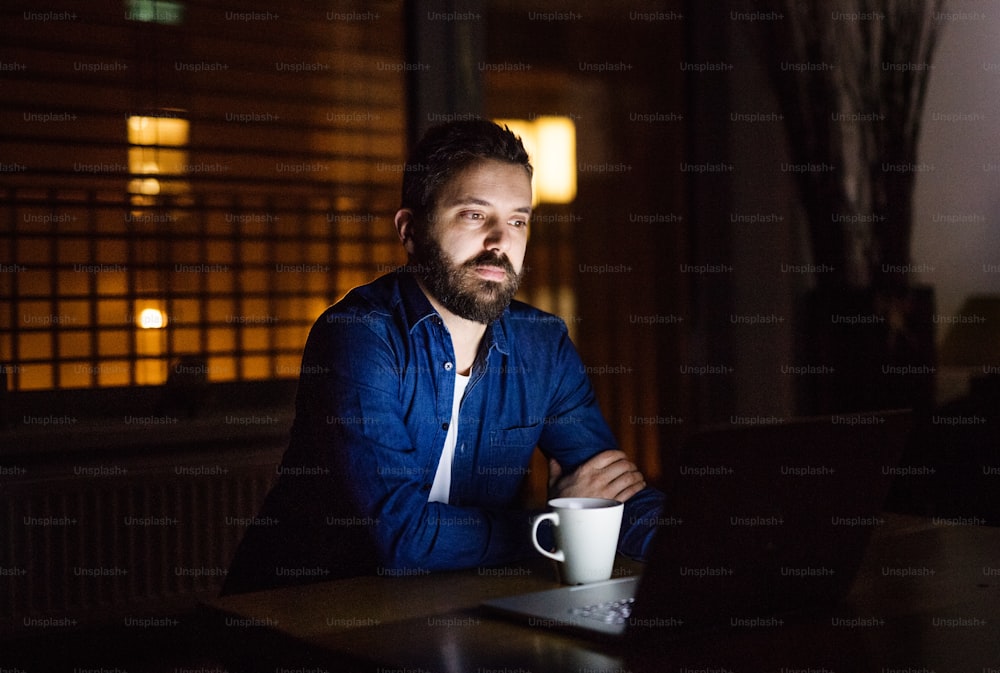 Ein gutaussehender Mann, der zu Hause oder nachts in einem Büro an einem Laptop arbeitet, mit einer Tasse Kaffee.