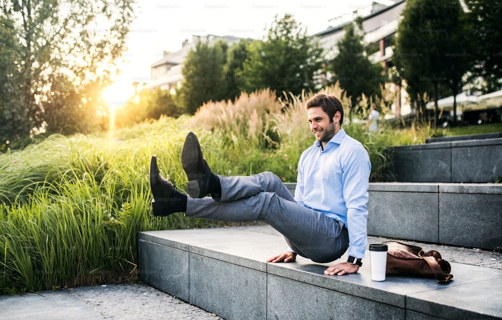 Un homme d’affaires heureux à l’extérieur sur des escaliers en béton au coucher du soleil, handstand en position L-assis.