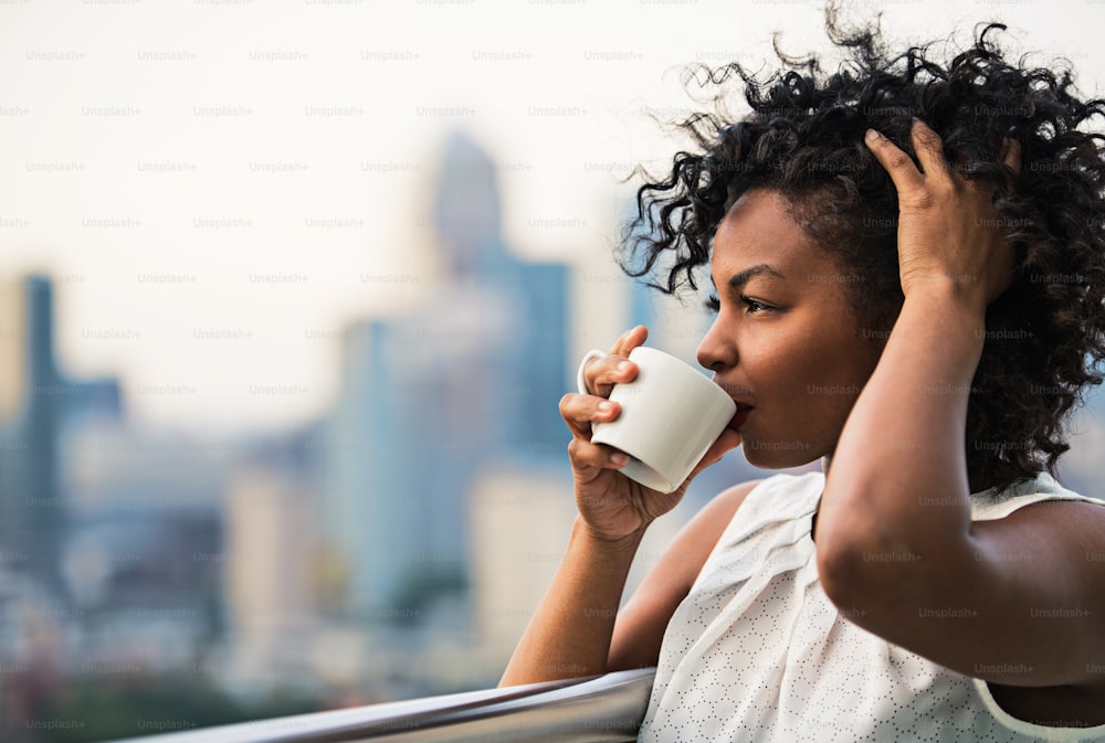 A close-up portrait of a black woman standing on a terrace, drinking coffee. Copy space.