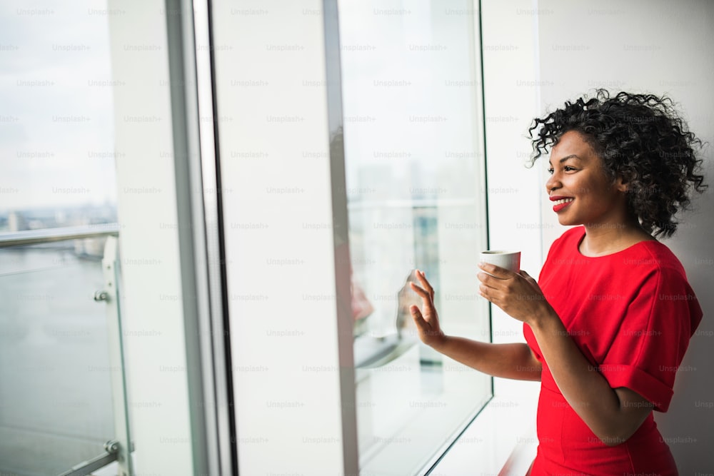 A happy black woman standing by the window holding coffee cup, looking out. Copy space.
